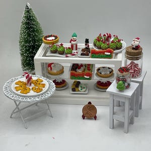 Puede incluir: Un expositor de panadería navideña en miniatura con un soporte blanco de varios niveles lleno de golosinas festivas, que incluyen galletas de jengibre, cupcakes, tartas y bastones de caramelo. Hay una pequeña mesa blanca con un plato de galletas y un árbol de Navidad en miniatura en un frasco.