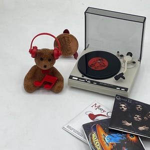 Könnte beinhalten: Ein Miniatur-Plattenspieler mit einem braunen Teddybär, der rote Kopfhörer trägt und eine Musiknote hält. Neben dem Player liegen vier Miniatur-Schallplatten, darunter "Queen II", "Frohe Weihnachten" und "Boston".