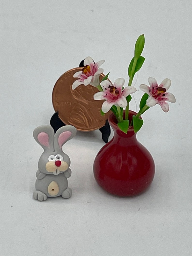 May include: Miniature gray bunny figurine with pink-lined ears and a red nose. A small red vase holds delicate white and pink flowers. A penny is used to show the scale of the tiny objects.