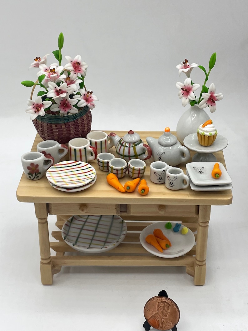 May include: Miniature wooden table with a collection of dollhouse accessories. Includes a basket of pink and white flowers, teacups, teapots, plates, carrots, and a cupcake. The table is made of light-colored wood.