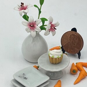 May include: Miniature Easter-themed arrangement featuring a white vase with pink-spotted lilies, a cupcake with a carrot on top, small carrots, and two square plates with a bunny design. A penny is displayed on a small stand.