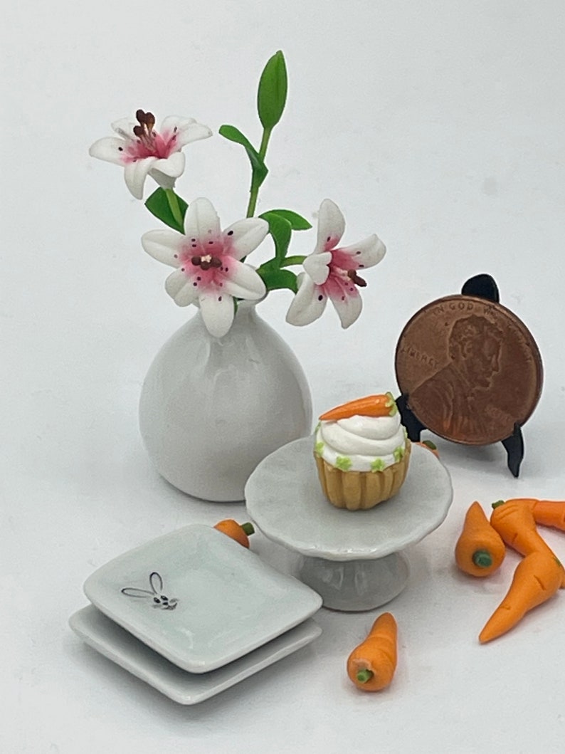 May include: Miniature Easter-themed arrangement featuring a white vase with pink-spotted lilies, a cupcake with a carrot on top, small carrots, and two square plates with a bunny design. A penny is displayed on a small stand.
