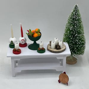Könnte beinhalten: Ein kleiner weißer Tisch mit zwei Ablagen, dekoriert mit einer Miniatur-Weihnachtsszene. Die Szene beinhaltet einen kleinen grünen Baum, eine Schale mit orangenen Früchten und mehrere weiße Kerzen auf einer Holzscheibe. Es gibt auch zwei kleine Kerzenhalter mit roten und grünen Sockeln.
