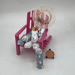 May include: A pink miniature beach chair with a white straw hat and rolled up beach towels with various patterns. A single rolled up towel with a seagull pattern is in the foreground.