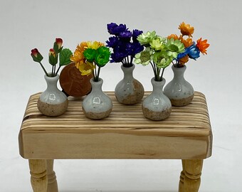 Puede incluir: Seis pequeños jarrones de cerámica con arreglos de flores artificiales de diferentes colores sobre una mesa de madera con patas torneadas. Los jarrones están moteados con esmalte marrón y blanco.