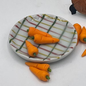 May include: A miniature ceramic plate with a white background and a plaid pattern in red, green, and yellow holds several small orange carrots with green stems. Additional carrots are scattered around the plate.