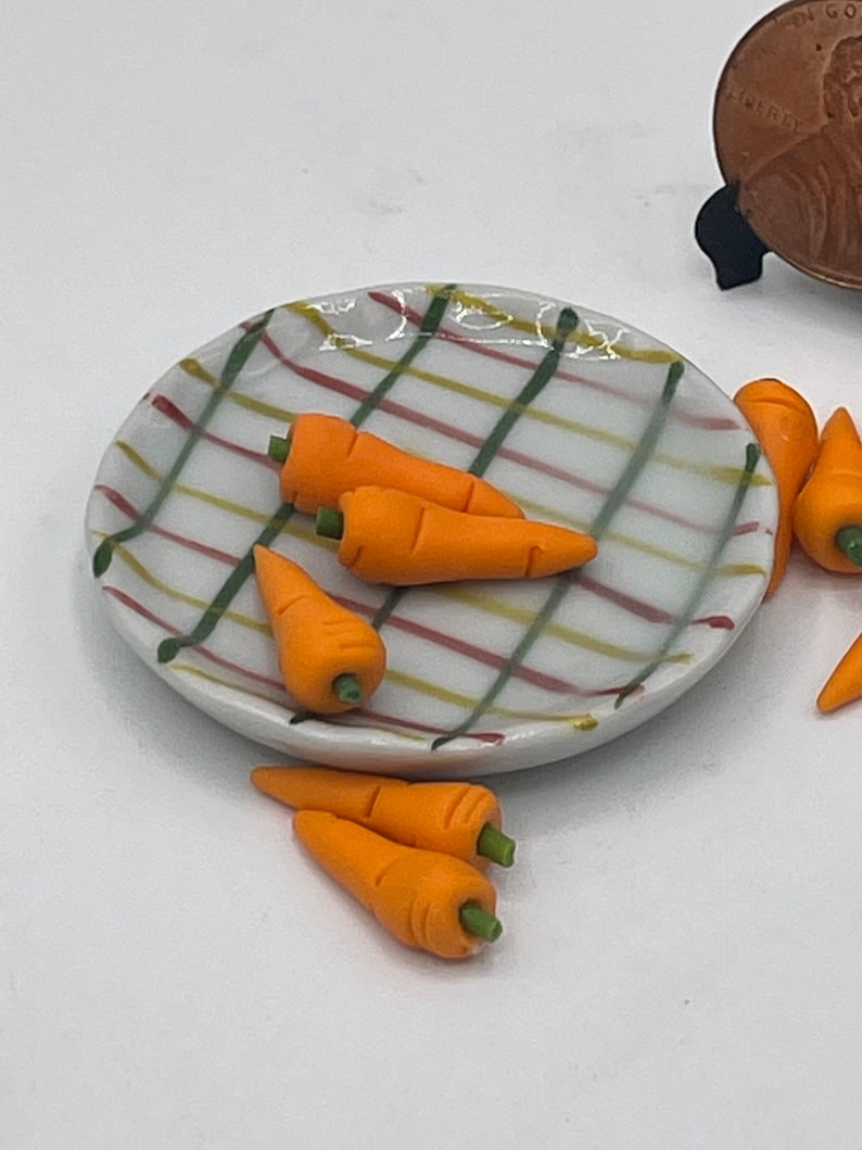 May include: A miniature ceramic plate with a white background and a plaid pattern in red, green, and yellow holds several small orange carrots with green stems. Additional carrots are scattered around the plate.