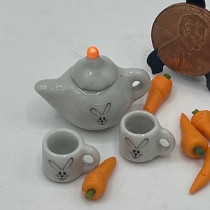 May include: A miniature gray ceramic tea set with a teapot, two cups, and several orange carrot figurines. Each piece of the tea set has a bunny face design. The teapot has an orange knob on the lid.