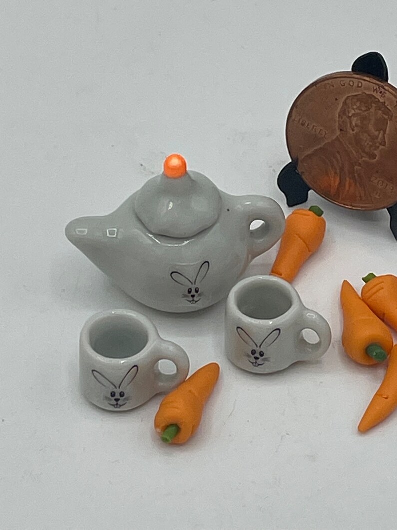 May include: A miniature gray ceramic tea set with a teapot, two cups, and several orange carrot figurines. Each piece of the tea set has a bunny face design. The teapot has an orange knob on the lid.