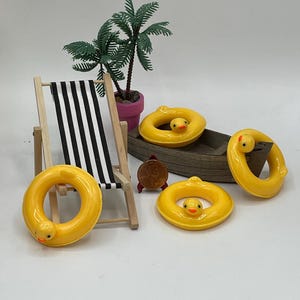 May include: A miniature beach scene with yellow duck-shaped pool floats, a black and white striped beach chair, a small boat, a potted palm tree, and a penny. The floats have a smiling duck face.
