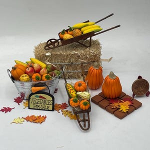 May include: A miniature fall harvest scene with a wheelbarrow filled with corn, pumpkins, and gourds. A second wheelbarrow holds more pumpkins and gourds. A basket overflowing with apples, pumpkins, and gourds sits next to a sign that reads "Vegetables for Sale".