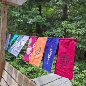 Peut inclure: Une guirlande de drapeaux rectangulaires en tissu suspendus sur une corde, présentant diverses couleurs dont le rouge, l'orange, le bleu et le violet. Les drapeaux affichent des motifs imprimés comme une coccinelle, un symbole om, du fromage et une tortue.