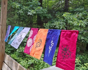 May include: A string of rectangular fabric flags hanging on a line, featuring various colors including red, orange, blue, and purple. The flags display printed designs such as a ladybug, an om symbol, a piece of cheese, floral patterns, and a turtle.