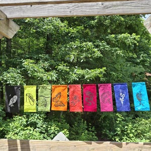 May include: A horizontal string of nine rectangular fabric banners in a rainbow gradient, each featuring a unique black or white screen-printed design including an owl, butterfly, chrysalis, dragon, fish, and the Om symbol, hanging outdoors.