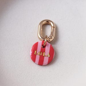May include: A round, red and pink striped charm with the name "LOLA" in gold lettering. The charm is attached to a gold-colored clasp. The charm is a pet tag or accessory.