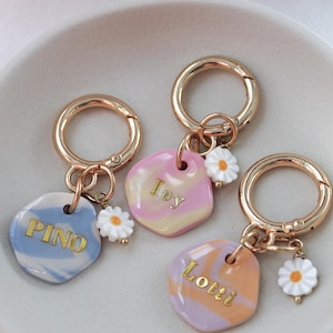 May include: Three personalised keychains with gold-coloured hardware and daisy charms. Each keychain features a unique, colourful, marbled tag with a name in gold lettering: "Pino", "Ivy", and "Lotti". The keychains are displayed in a white dish.