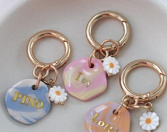 May include: Three personalized keychains with gold-colored hardware and daisy charms. Each keychain features a unique, colorful, marbled tag with a name in gold lettering: "Pino", "Ivy", and "Lotti". The keychains are displayed in a white dish.
