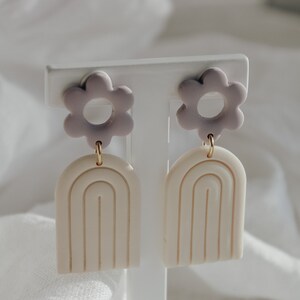 May include: A pair of earrings with a white arch design and a purple flower charm. The earrings are on a white display stand.