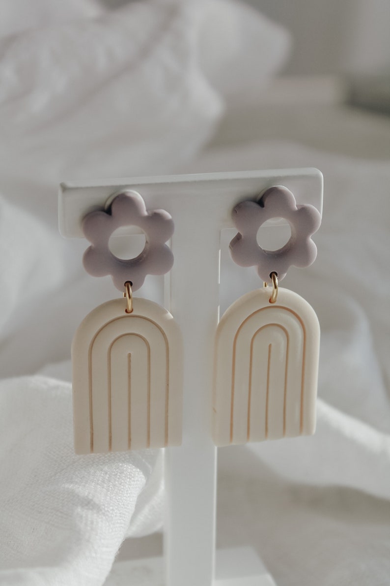 May include: A pair of earrings with a white arch design and a purple flower charm. The earrings are on a white display stand.
