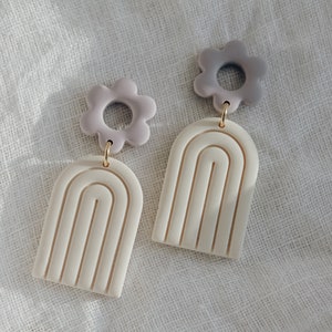 May include: A pair of earrings with a white arch shape and a purple flower charm. The earrings are made of polymer clay.