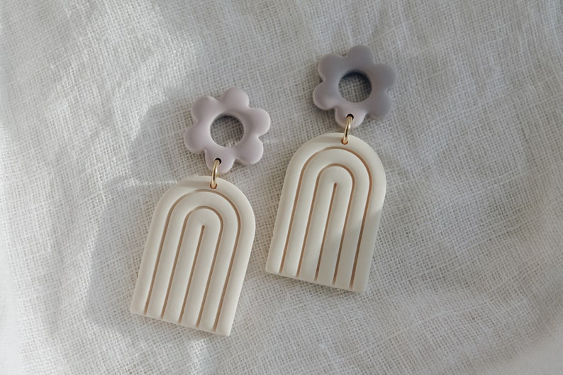 May include: A pair of earrings with a white arch shape and a purple flower charm. The earrings are made of polymer clay.