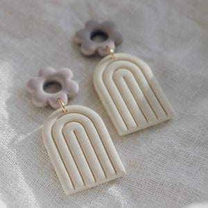 May include: A pair of handmade polymer clay earrings featuring a white arch shape with a textured design and a light purple flower charm.