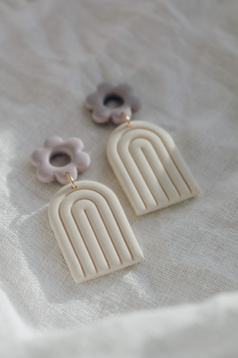 May include: A pair of handmade polymer clay earrings featuring a white arch shape with a textured design and a light purple flower charm.