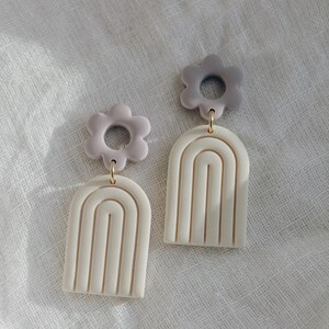 May include: A pair of white and purple polymer clay earrings. Each earring features a white arch with a raised design and a purple flower charm.
