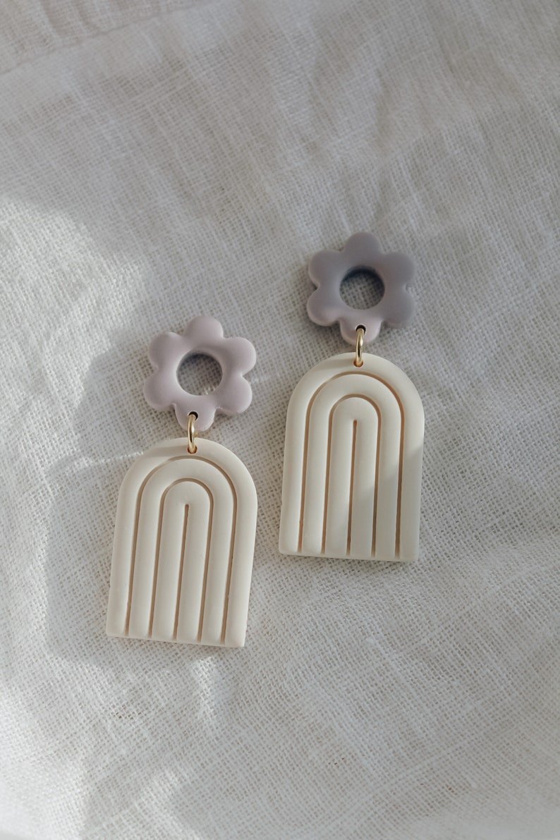 May include: A pair of white and purple polymer clay earrings. Each earring features a white arch with a raised design and a purple flower charm.