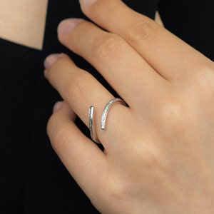 Pu&ograve; includere: Un anello argentato dal design avvolgente. Un lato &egrave; una barra dritta, l'altro si curva attorno al dito, impreziosito da piccole pietre scintillanti. L'anello &egrave; mostrato su una mano su sfondo scuro.