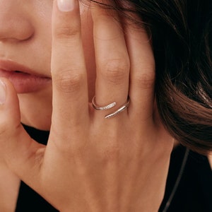 Pu&ograve; includere: Un anello color argento con un design avvolgente. L'anello presenta piccole pietre scintillanti lungo un lato. Il gioiello &egrave; indossato su un dito, con la mano e il braccio visibili su uno sfondo neutro.