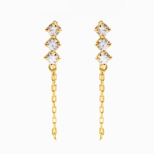 May include: A pair of gold earrings featuring three clear gemstones in a vertical arrangement. Each earring has a fine chain pendant. The earrings are displayed against a plain white backdrop, highlighting their design.