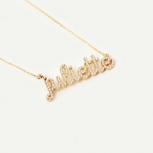 Könnte beinhalten: Eine goldene Halskette mit dem Namen "Juliette" in kursiv geschriebener Schrift. Die Buchstaben sind mit kleinen, klaren Edelsteinen umrandet.