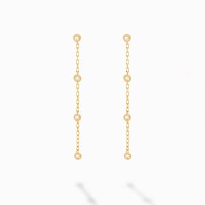 May include: A pair of gold drop earrings featuring a delicate chain design. Each earring consists of a thin gold chain interspersed with four small, round, clear stones set in gold bezels, creating a vertical, linear, and dangling appearance.
