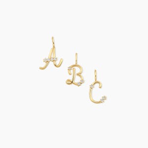 May include: Three gold-coloured initial pendants, the letters A, B, and C, each adorned with small, sparkling gemstones. The pendants are in a cursive font and are displayed against a white background.