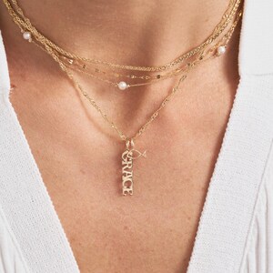 Pode incluir: Um colar de ouro com um pingente que diz "GRACE" e um pequeno charme de peixe. O colar &eacute; sobreposto com outras correntes de ouro e detalhes de p&eacute;rolas.