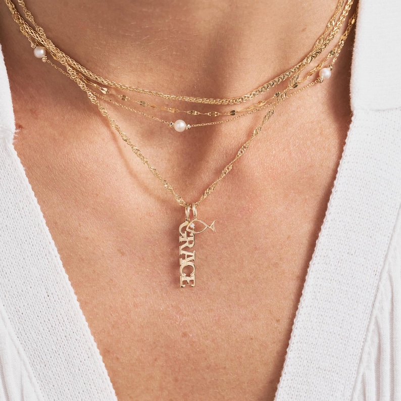 Pode incluir: Um colar de ouro com um pingente que diz "GRACE" e um pequeno charme de peixe. O colar &eacute; sobreposto com outras correntes de ouro e detalhes de p&eacute;rolas.