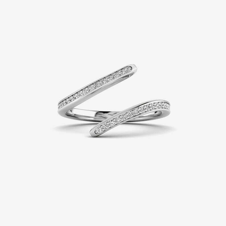 Pu&ograve; includere: Un anello argentato con un design aperto unico. L'anello presenta due fasce curve, ciascuna ornata di piccole pietre chiare e scintillanti. Le fasce si incrociano con grazia, creando un look elegante e moderno. L'anello &egrave; posto su uno sfondo bianco.