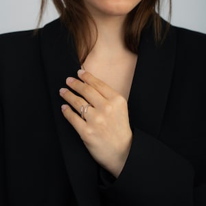 Pu&ograve; includere: Un anello color argento dal design minimalista. L'anello avvolge il dito con una curva sottile ed &egrave; adornato da piccole pietre scintillanti. L'anello &egrave; mostrato su una mano, su uno sfondo di blazer nero.