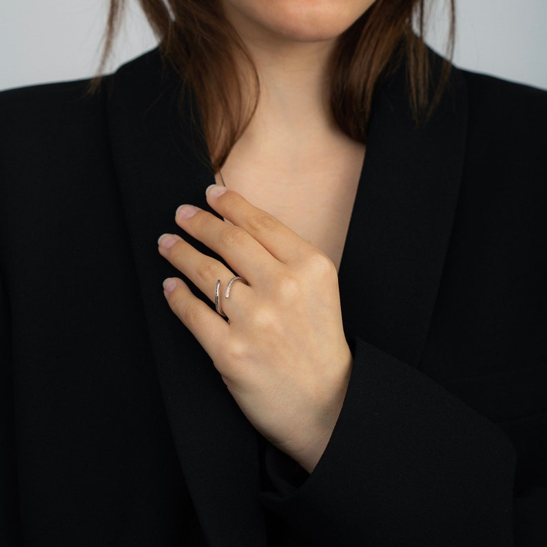 Pu&ograve; includere: Un anello color argento dal design minimalista. L'anello avvolge il dito con una curva sottile ed &egrave; adornato da piccole pietre scintillanti. L'anello &egrave; mostrato su una mano, su uno sfondo di blazer nero.