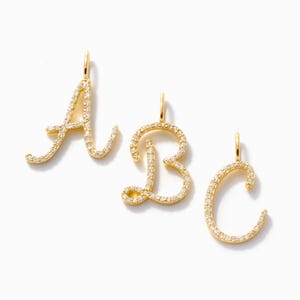 Könnte beinhalten: Drei goldfarbene Anhänger mit kursiven Buchstaben: A, B und C. Jeder Buchstabe ist mit kleinen, klaren Edelsteinen besetzt. Die Anhänger haben oben eine Schlaufe zum Befestigen an einer Kette oder einem Armband. Die Buchstaben werden auf weißem Hintergrund angezeigt.