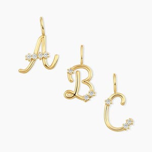 May include: Three gold letter pendants in a cursive script font, specifically the letters A, B, and C. Each pendant features small, clear, round-cut stones embedded along the gold metal surface, with a small loop at the top for attachment.