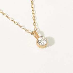 May include: A gold necklace featuring a delicate chain and a single round, clear gemstone set in a polished gold bezel setting. The pendant hangs from a small, solid gold bail, showcasing a clean, reflective surface and a classic, simple design.