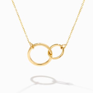 May include: A gold-toned necklace featuring a delicate chain and a pendant composed of two interlocking circular rings of different sizes. The metal has a polished, reflective finish, and the design is suspended from a thin, matching link chain.