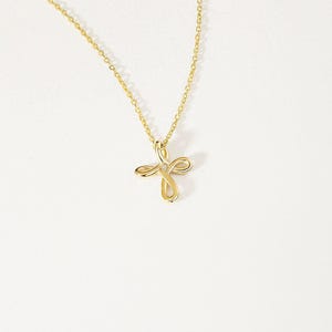 May include: A gold necklace featuring a delicate chain and a pendant shaped like a stylized cross with looped, ribbon-like arms. A single small, clear gemstone is set at the center where the four loops of the cross meet, creating a polished finish.