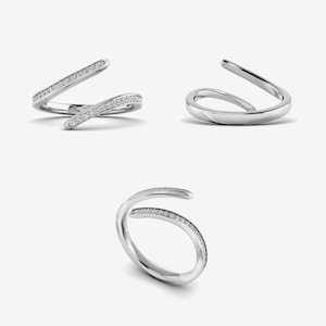 Pu&ograve; includere: Tre immagini di un anello d'argento dal design unico e aperto. Un anello presenta una fascia foderata con piccole pietre trasparenti. Gli altri due anelli hanno una superficie liscia e lucida. Il design dell'anello si curva e si sovrappone, creando un look elegante e moderno. Gioielli.