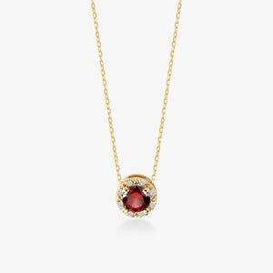 May include: A gold chain necklace featuring a circular pendant with a round, deep red gemstone at the center. The red stone is surrounded by a halo of small, clear, sparkling stones set in a gold frame, suspended from a delicate gold link chain.