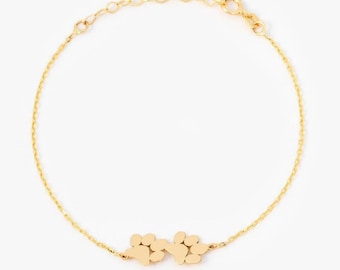 May include: A gold-toned chain bracelet featuring two connected paw print charms at the center. The bracelet is constructed with delicate circular links and includes a small lobster claw clasp mechanism at the end of the chain for secure fastening.