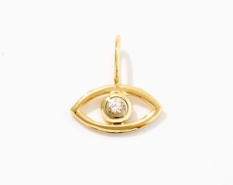 May include: A gold pendant shaped like an eye with a polished finish. The center of the eye features a small, round, clear gemstone set within a circular frame. A small gold jump ring is attached to the top of the eye-shaped pendant for hanging.