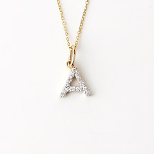 May include: A gold chain necklace featuring a letter A pendant. The pendant is encrusted with small, clear, round-cut stones along its front surface. The chain is thin and delicate, connecting to the top of the letter via a small gold jump ring.